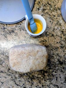 Chilled spiced hazelnut Linzertorte dough block wrapped in plastic wrap beside beaten egg yolk and pastry brush