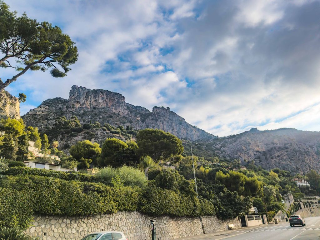 Steep limestone mountain cliffs towering over a quiet coastal road lined with pine trees above Eze sur Mer