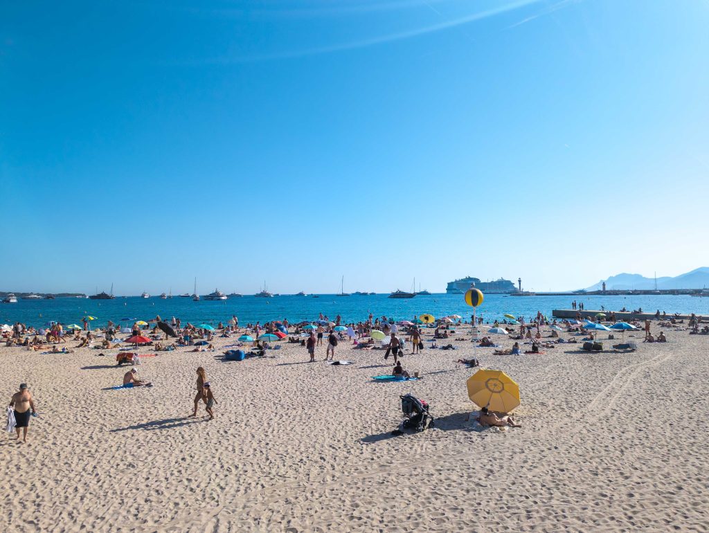 Crowded sandy beach in Cannes filled with sunbathers and colorful umbrellas overlooking yachts and a cruise ship anchored in the bay