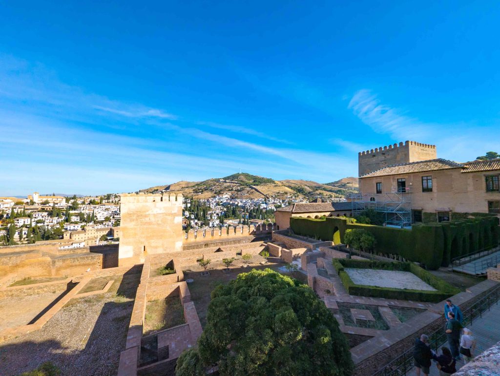 View across the brick ruins and manicured hedges of the Alhambra toward stone watchtowers and the whitewashed Albaicin hillside in Granada
