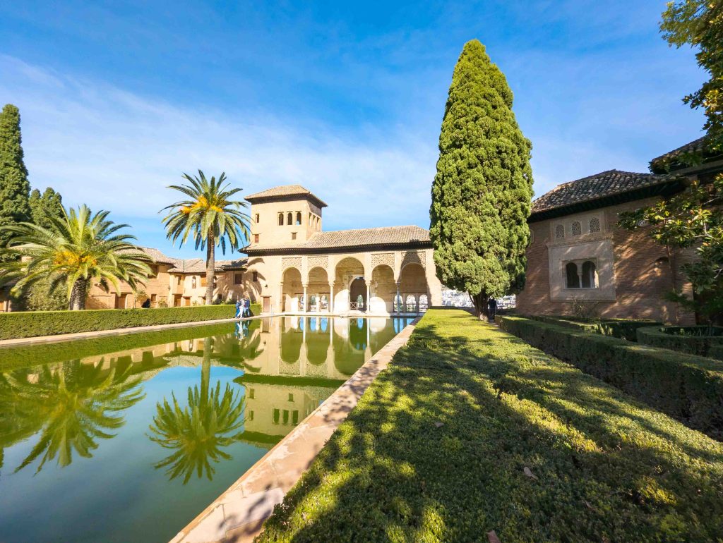 Arched portico of the Partal Palace reflected in a tranquil pool framed by a tall cypress tree and date palms at the Alhambra