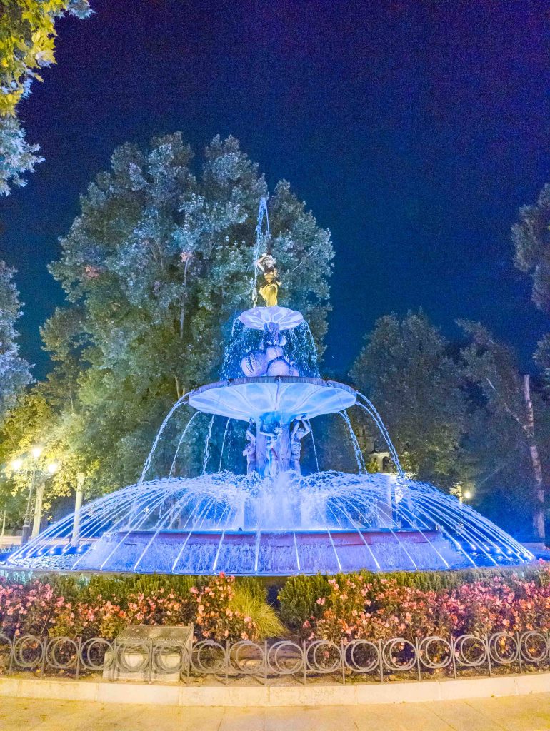 Tiered stone fountain of Fuente de las Granadas glowing with blue and gold illumination at night on a tree lined square in Granada