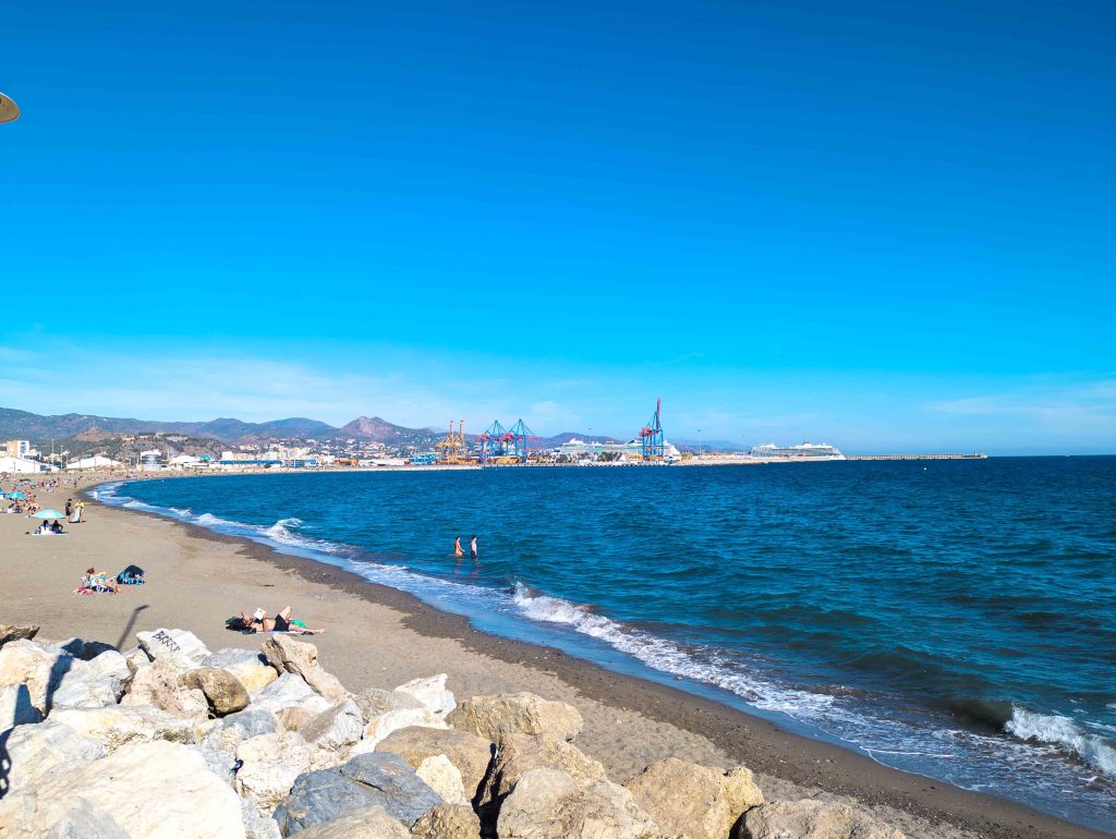 Sandy shoreline and gentle Mediterranean waves along Malagueta Beach in Malaga with port cranes and cruise ships on the horizon