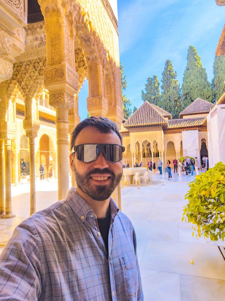 A traveler wearing sunglasses standing in the sunlit Court of the Lions surrounded by carved stucco arches and marble columns at the Alhambra